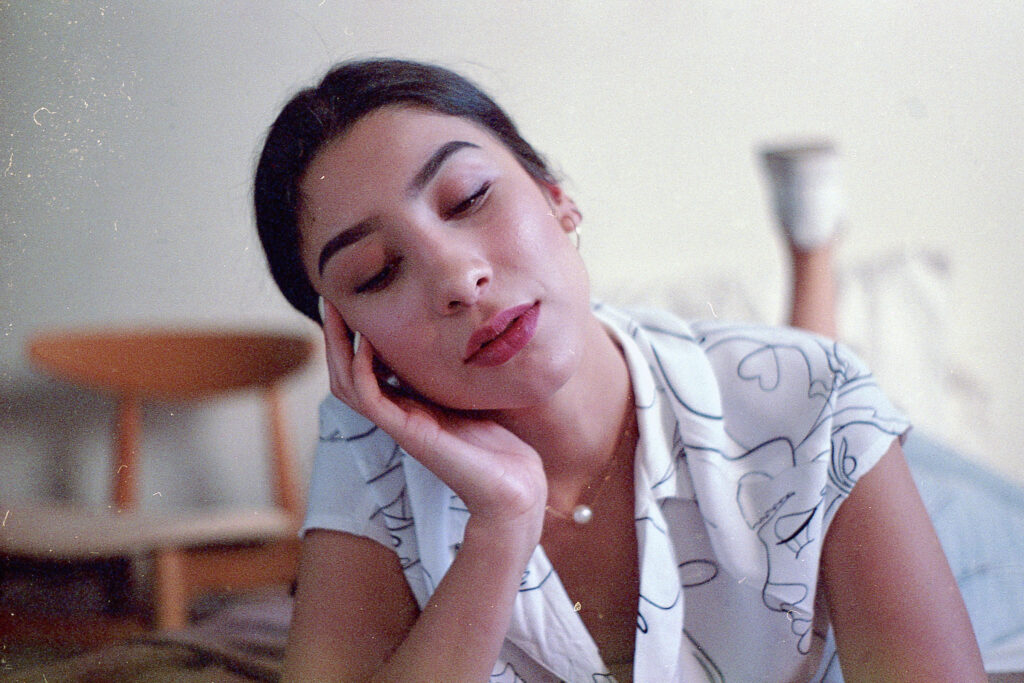 Nadya Inzunza Film Photo. A serene moment captured of a person with closed eyes resting their head on their hand wearing a white blouse with abstract patterns.